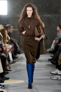 Model walks a fashion runway in a brown hoodie and matching skirt, bright blue tights, burgundy shoes, and a tan crossbody bag held at her side.
