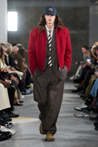 Male model on a runway wearing a red zip-front jacket over a brown plaid suit, white shirt, striped tie, and tan shoes.