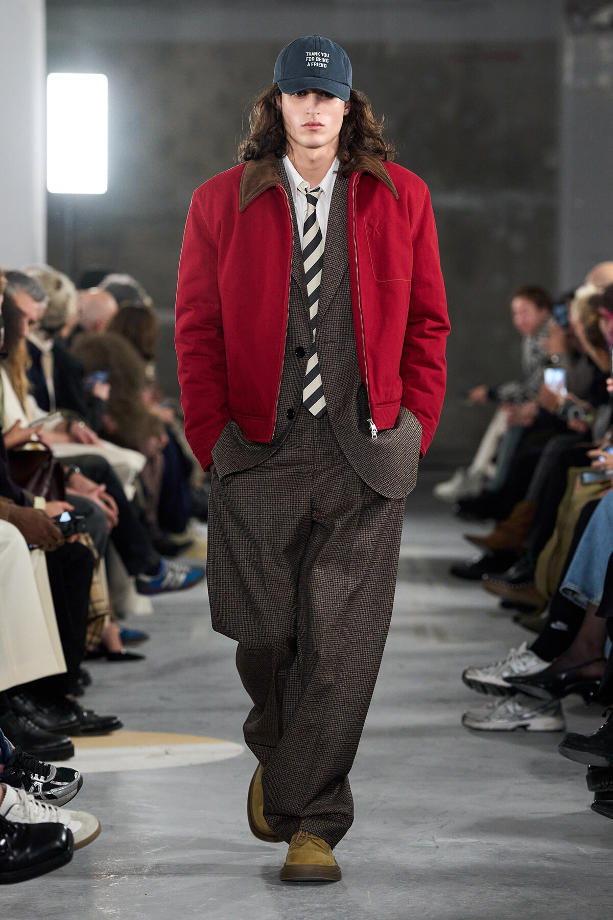 Male model on a runway wearing a red zip-front jacket over a brown plaid suit, white shirt, striped tie, and tan shoes.