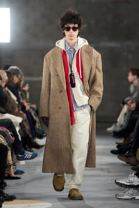 Male model walks the runway in a tan herringbone overcoat, striped shirt, white hoodie, red scarf, and wide white pants with sunglasses.