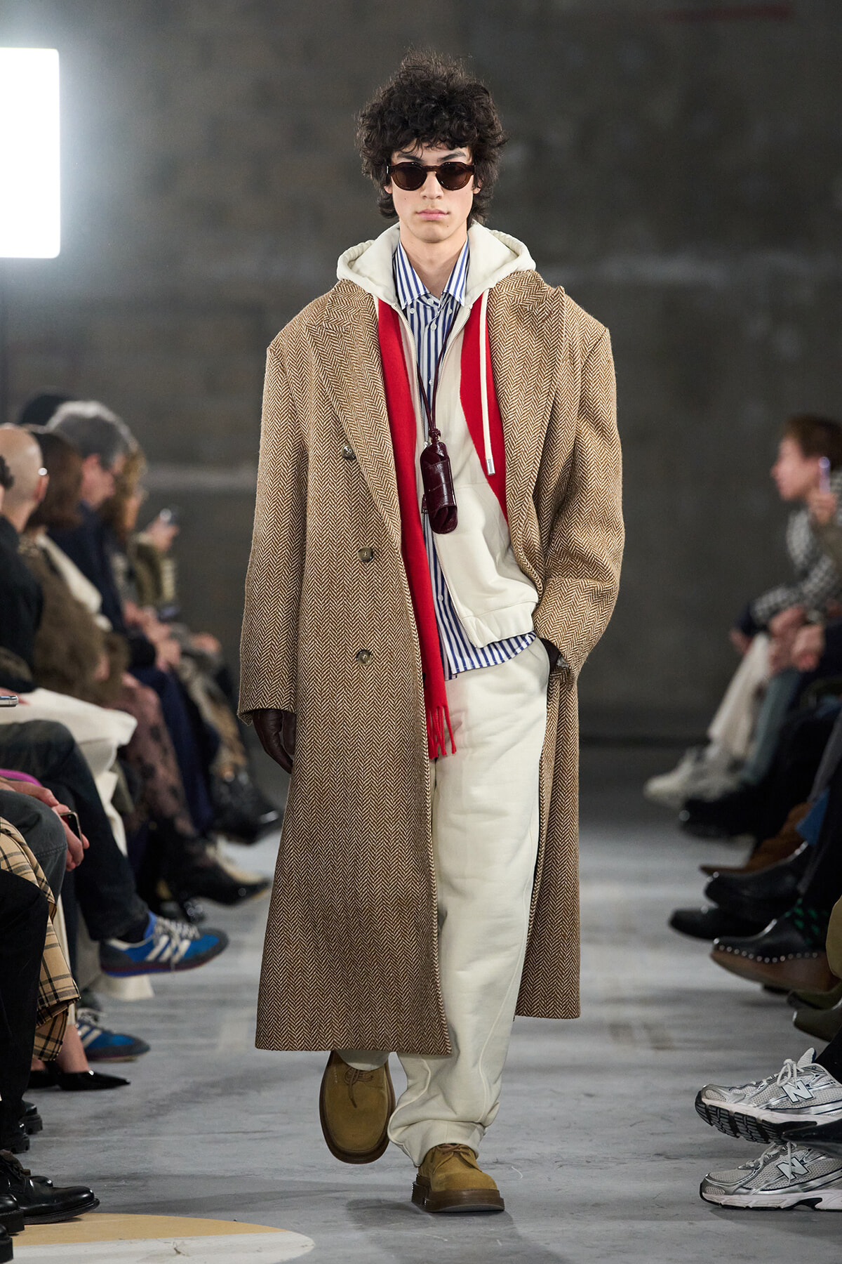 Male model walks the runway in a tan herringbone overcoat, striped shirt, white hoodie, red scarf, and wide white pants with sunglasses.
