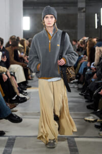 Model walks the runway in a gray fleece with mustard trim, loose beige wide-leg pants, a gray beanie, and a leopard-print bag.