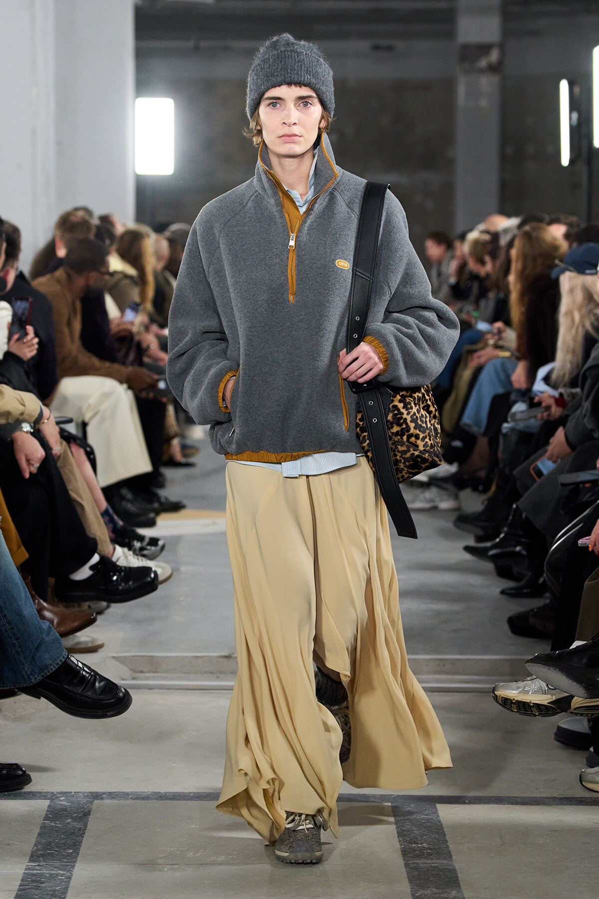 Model walks the runway in a gray fleece with mustard trim, loose beige wide-leg pants, a gray beanie, and a leopard-print bag.
