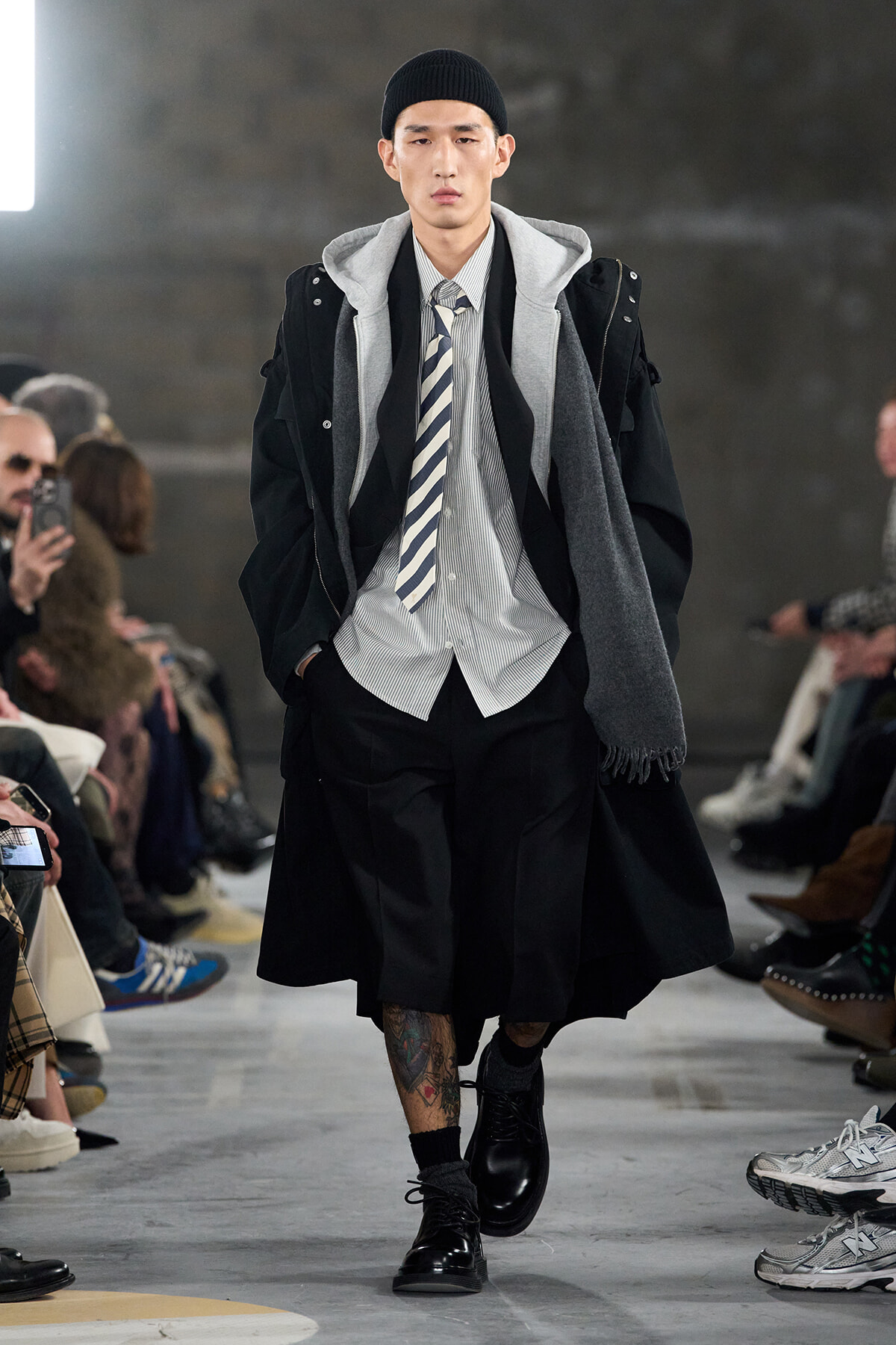 Male model walking a fashion runway in a layered streetwear outfit: black coat, gray hoodie, striped shirt and tie, black shorts, beanie, and tattoos visible on his lower leg.