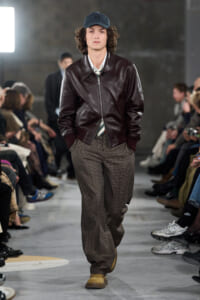 Male model walks a runway in a burgundy leather bomber, striped shirt, loose plaid trousers, tan boots, and a dark cap.