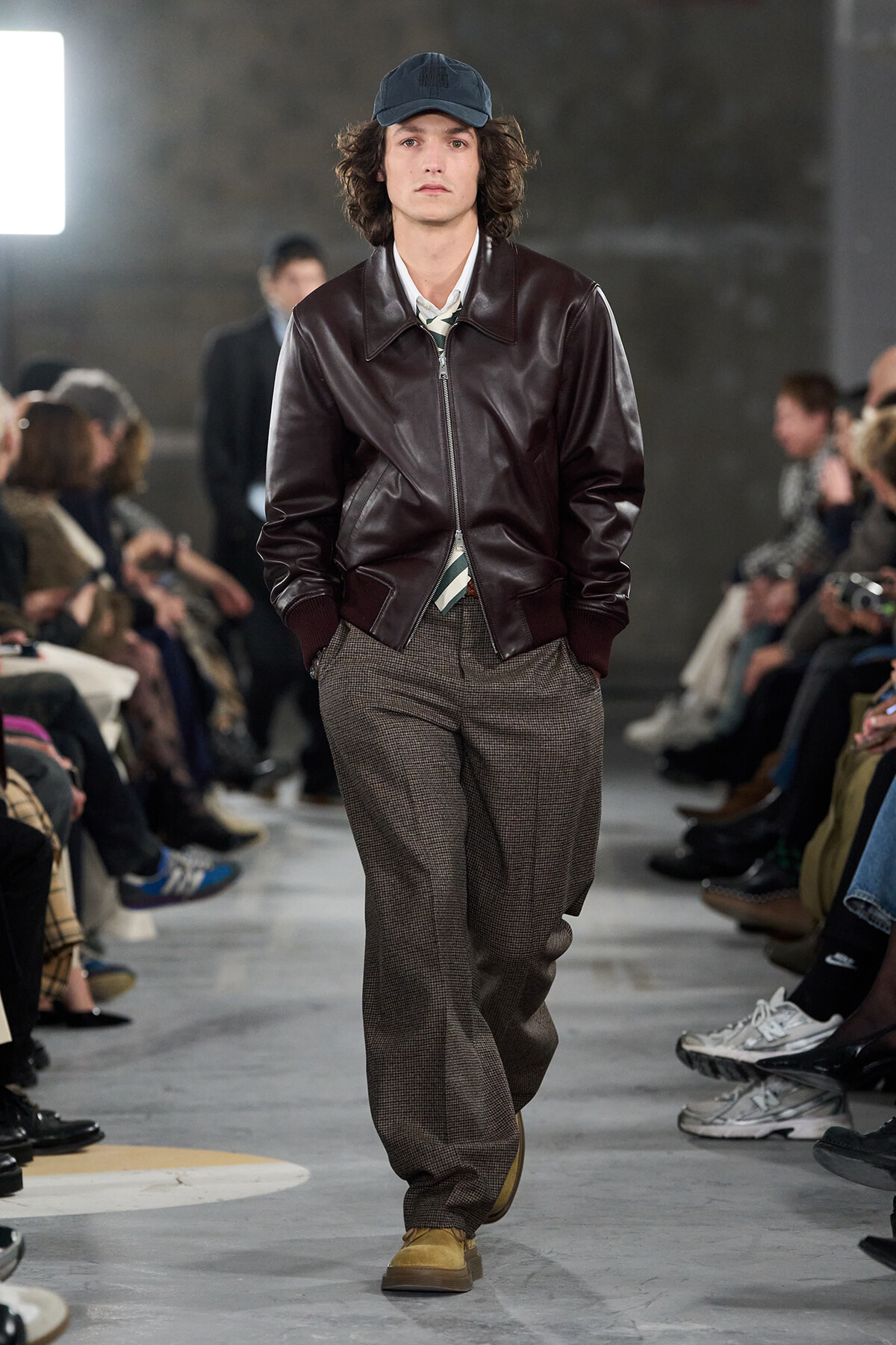 Male model walks a runway in a burgundy leather bomber, striped shirt, loose plaid trousers, tan boots, and a dark cap.