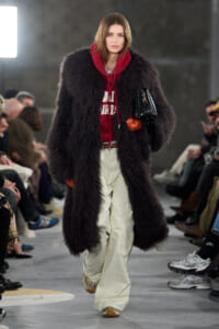 Model on a runway wearing a long black shaggy faux-fur coat over a red hoodie, cream pants, orange gloves, and a black handbag.