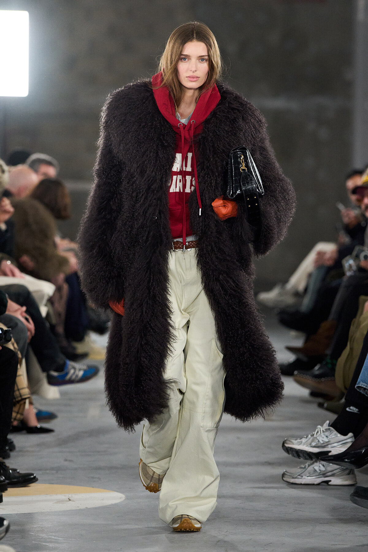 Model on a runway wearing a long black shaggy faux-fur coat over a red hoodie, cream pants, orange gloves, and a black handbag.