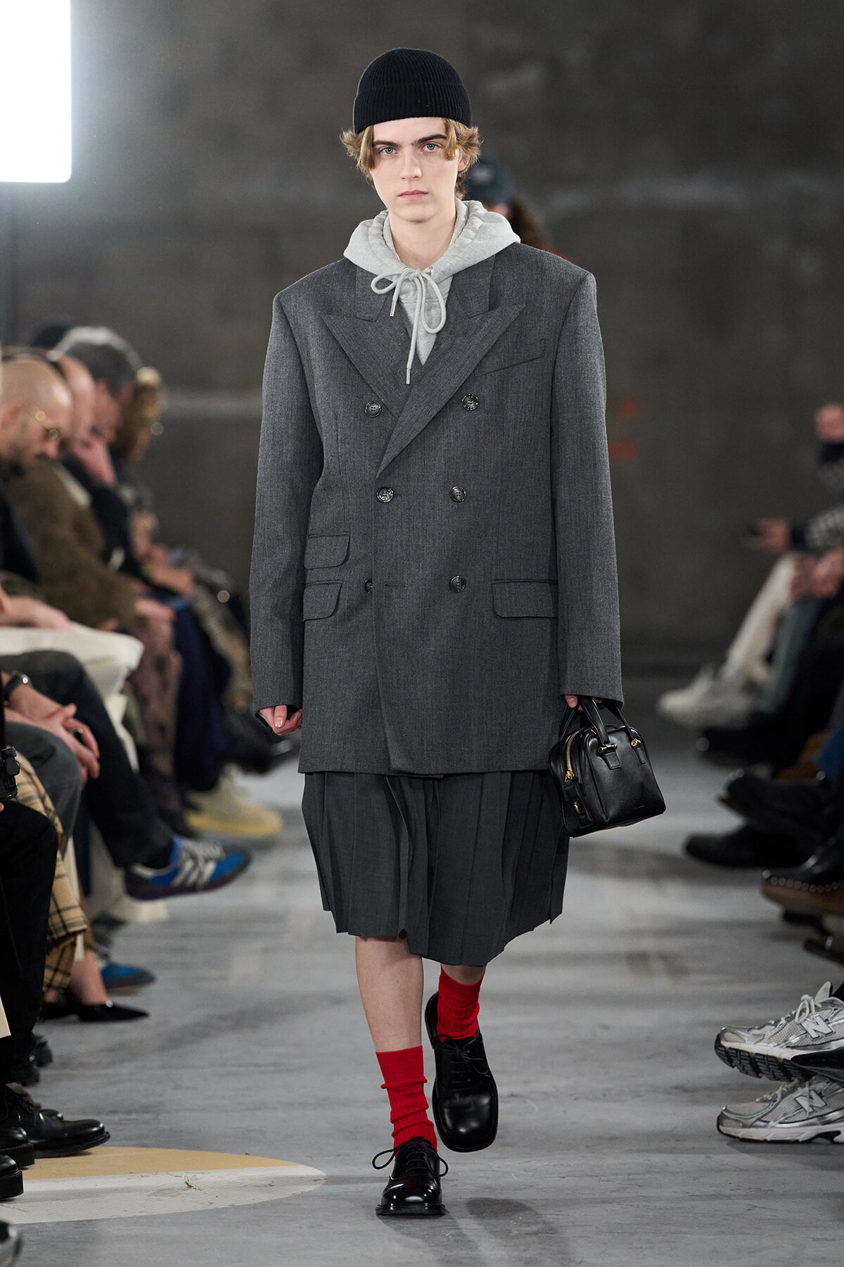 Male model walking a runway in a gray double-breasted blazer over a gray hoodie, red socks, black shoes, and a small black handbag.