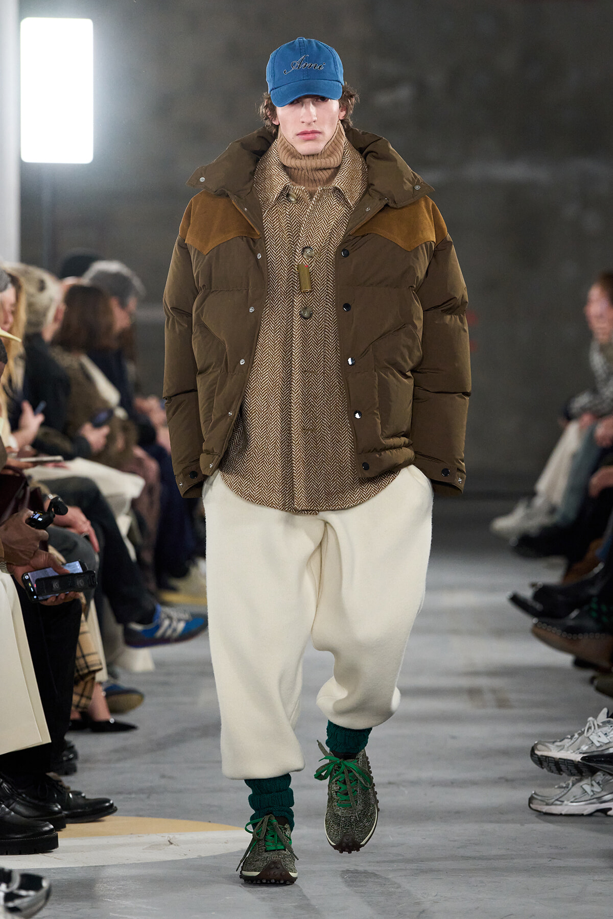 Runway model in a brown puffer jacket over a tan turtleneck and herringbone shirt, cream wide pants, green socks, and sneakers, wearing a blue cap named 'Anne'