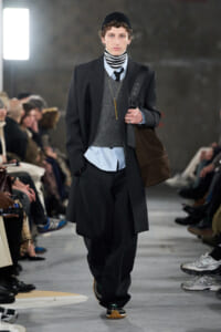 Male runway model in a dark overcoat, layered with a gray sweater and striped turtleneck, wearing a black beanie and carrying a brown bag on the shoulder.