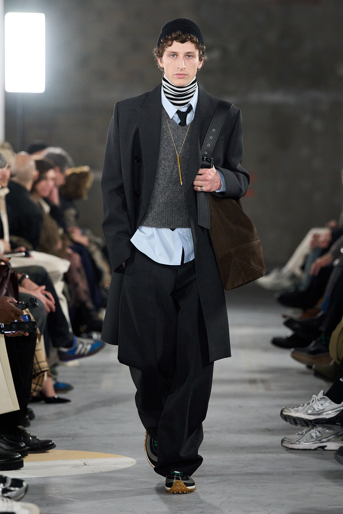 Male runway model in a dark overcoat, layered with a gray sweater and striped turtleneck, wearing a black beanie and carrying a brown bag on the shoulder.