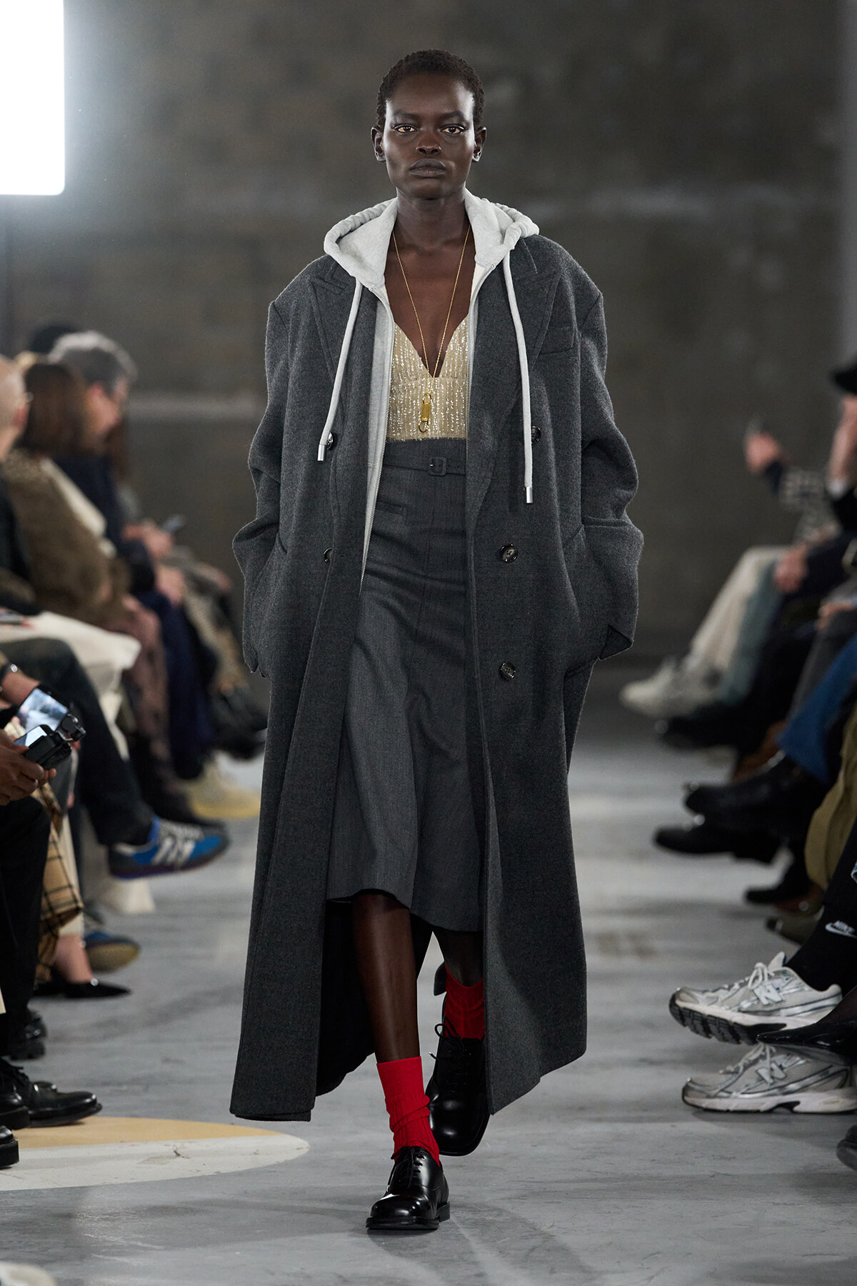 Model walking a fashion show in a long gray coat over a white hoodie, gold sequined top, and gray skirt, paired with bright red socks and black shoes.