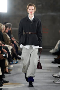 Model on a runway wearing a black belted oversized jacket over a white shirt, light gray baggy pants, blue leg warmers, and a burgundy handbag.