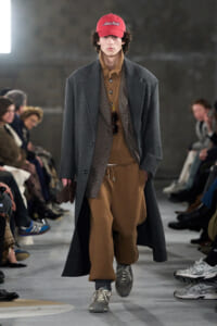 Male model walking a runway in a long gray overcoat over brown layered outfit with a red cap and sneakers, audience in background.
