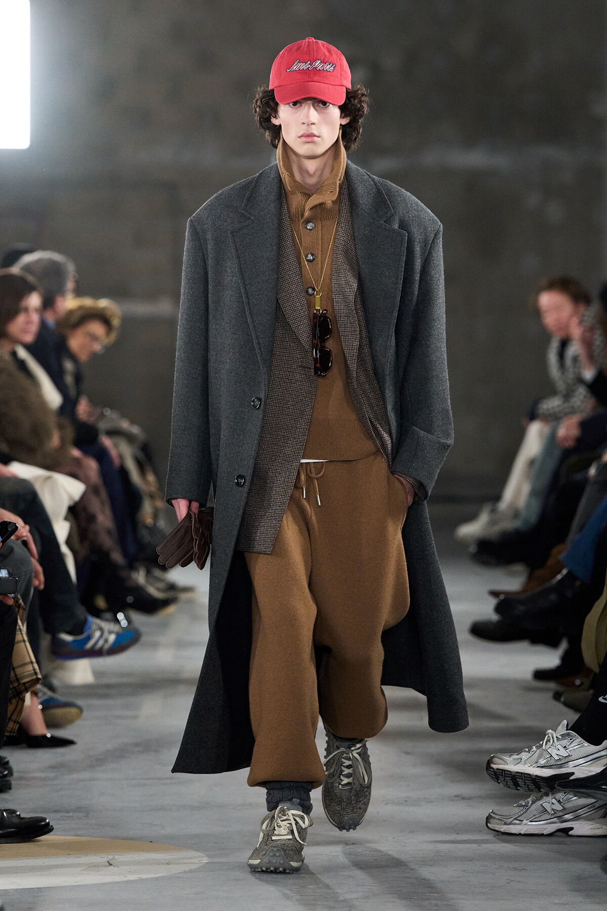 Male model walking a runway in a long gray overcoat over brown layered outfit with a red cap and sneakers, audience in background.