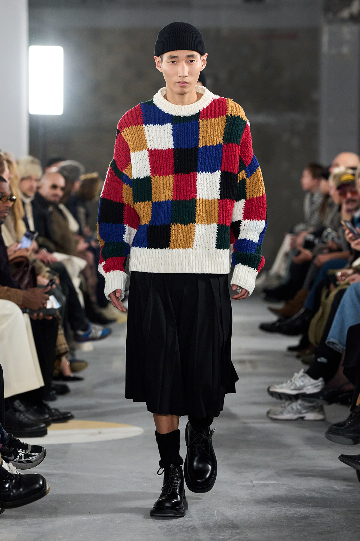 Male model walks a runway in a multicolor patchwork knit sweater, black beanie, wide black skirt, and combat boots.