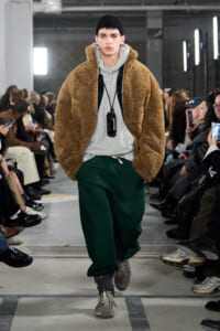 Male model walks the runway in a tan teddy-bear fleece jacket over a gray hoodie, green joggers, and gray sneakers with a black beanie.