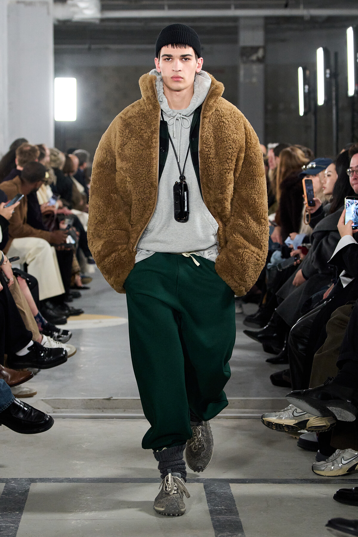 Male model walks the runway in a tan teddy-bear fleece jacket over a gray hoodie, green joggers, and gray sneakers with a black beanie.