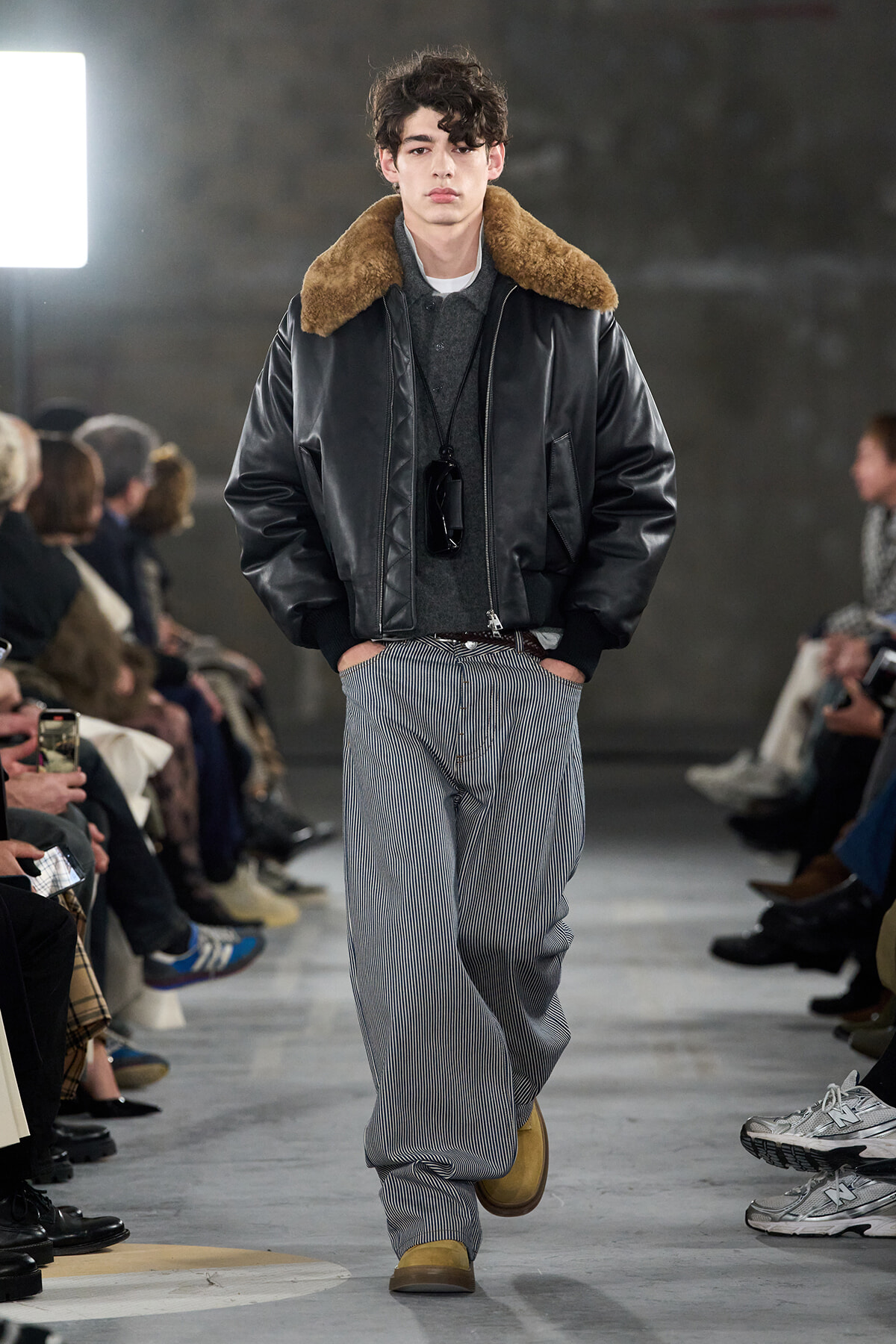 Male model on a runway wearing a black leather bomber with a brown shearling collar, gray knit top, and blue-pinstriped wide-leg pants, tan boots.
