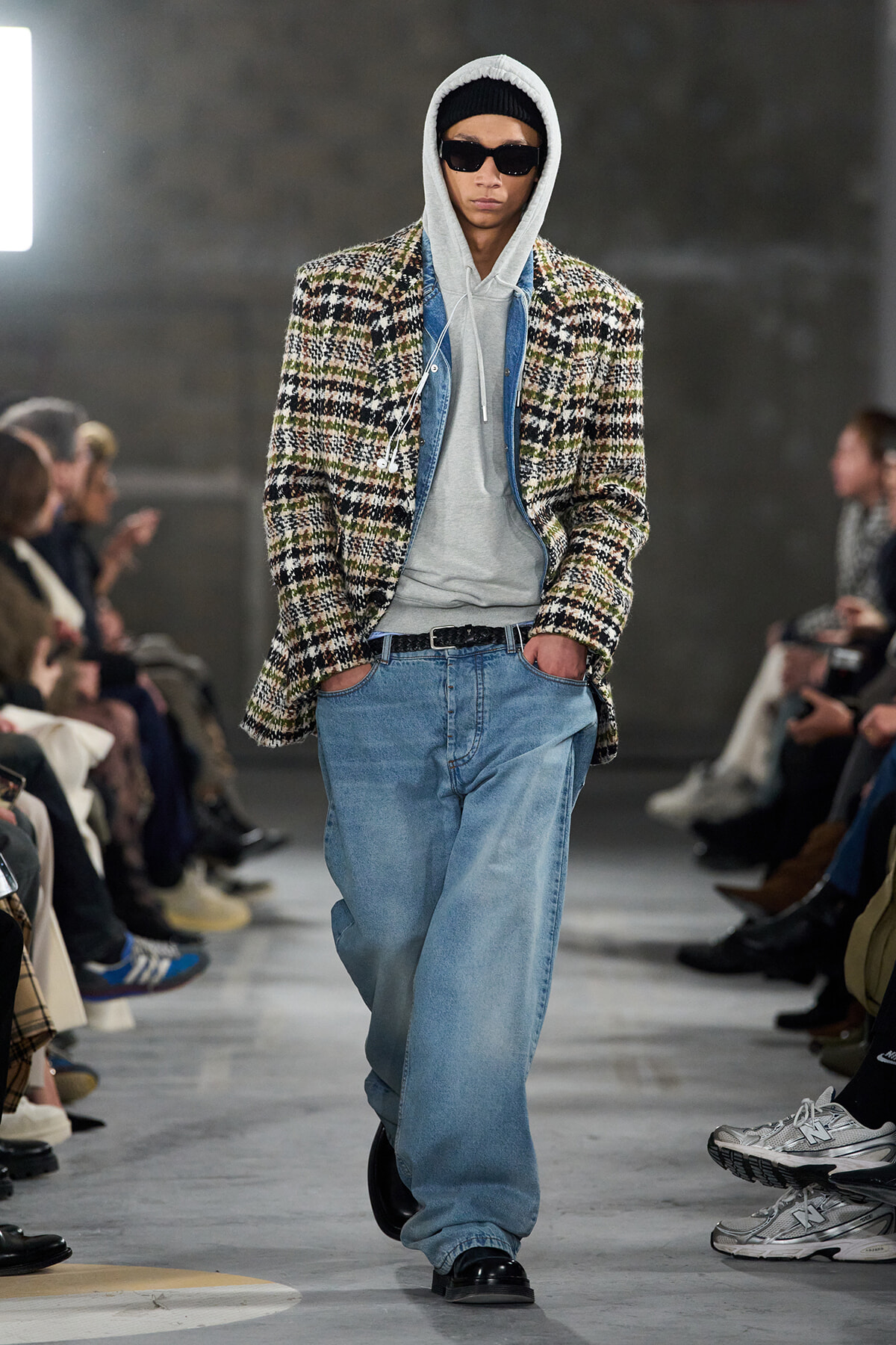 Male runway model wearing a plaid oversized coat over a gray hoodie and light-wash jeans, with black sunglasses and a knit cap, walking the catwalk.