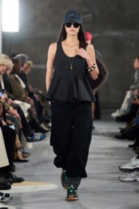 Model walks a runway in an all-black outfit: sleeveless peplum top, wide-leg pants, cap, sunglasses, and layered necklaces.