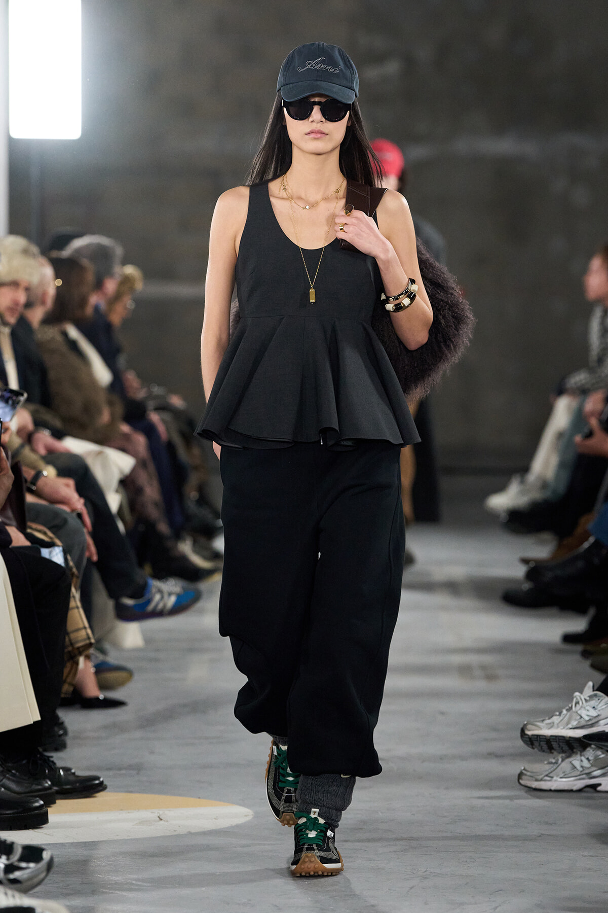 Model walks a runway in an all-black outfit: sleeveless peplum top, wide-leg pants, cap, sunglasses, and layered necklaces.