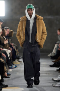 Male model on a fashion runway wearing a tan shearling jacket over a gray hoodie and dark denim overalls, with a green cap, walking past seated audience.