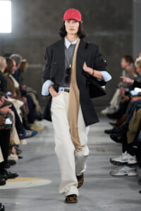 Model on a fashion runway wearing an oversized black blazer, light blue shirt, gray sweater, beige scarf, white wide‑leg pants, leopard print shoes, and a red cap.