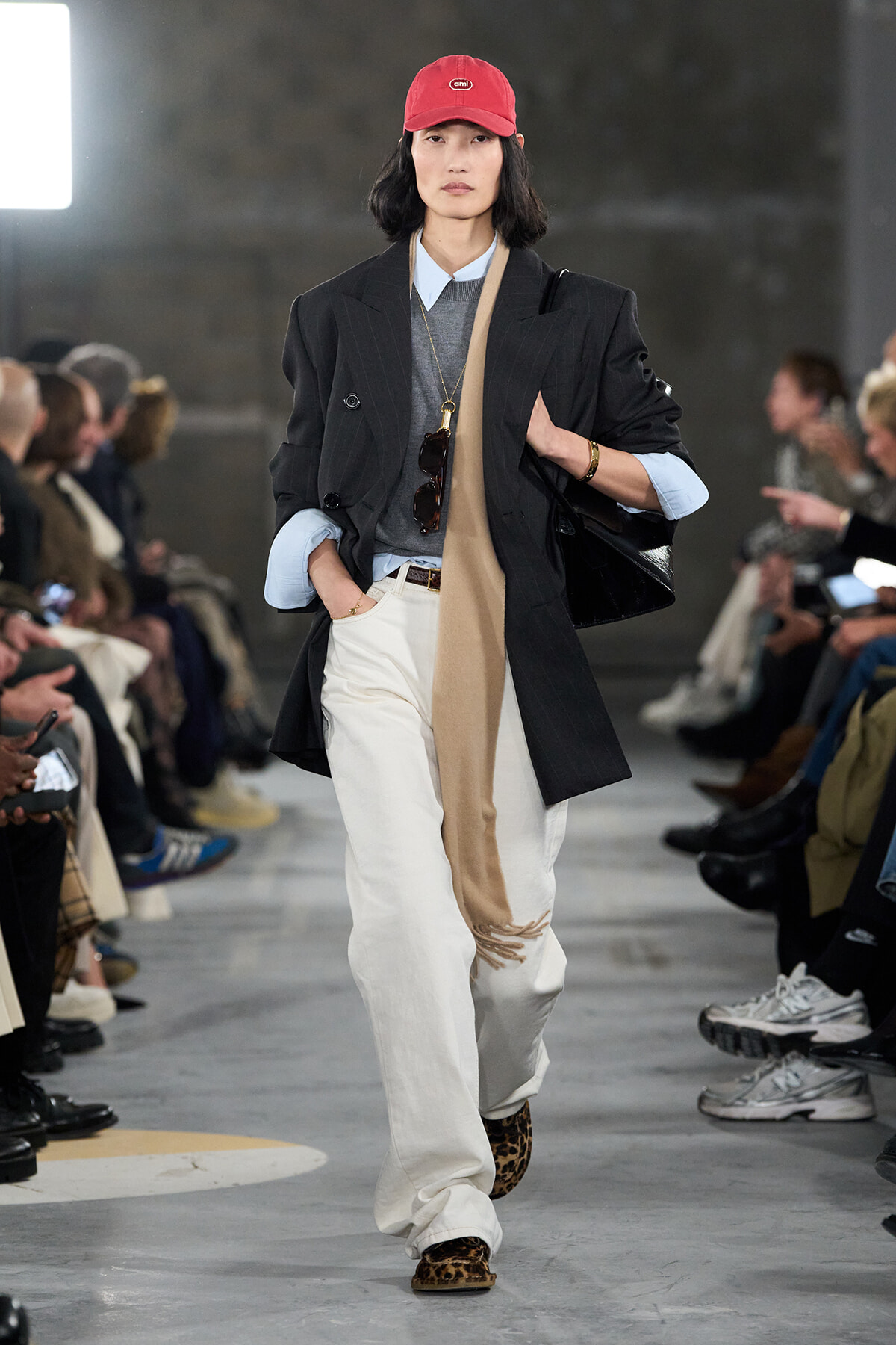 Model on a fashion runway wearing an oversized black blazer, light blue shirt, gray sweater, beige scarf, white wide‑leg pants, leopard print shoes, and a red cap.