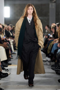 Male model walking a fashion show runway in a tan trench coat over a dark pinstripe suit with a white shirt and striped tie, audience watching on both sides.