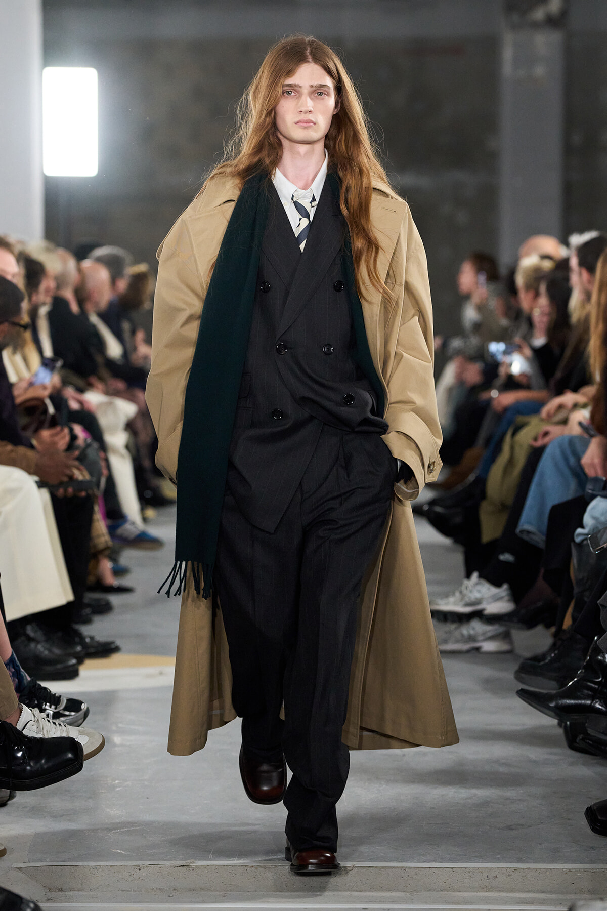 Male model walking a fashion show runway in a tan trench coat over a dark pinstripe suit with a white shirt and striped tie, audience watching on both sides.
