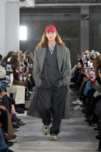 Model walks the runway in a gray three-piece suit with a long herringbone coat and red cap, as photographers film from the sides.