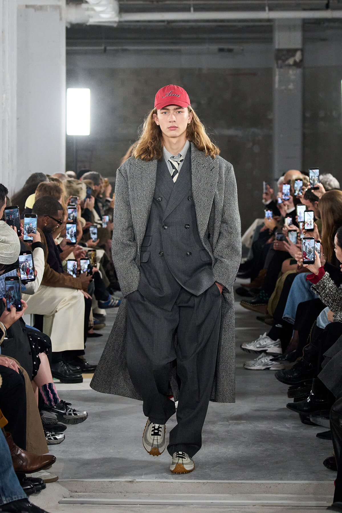 Model walks the runway in a gray three-piece suit with a long herringbone coat and red cap, as photographers film from the sides.