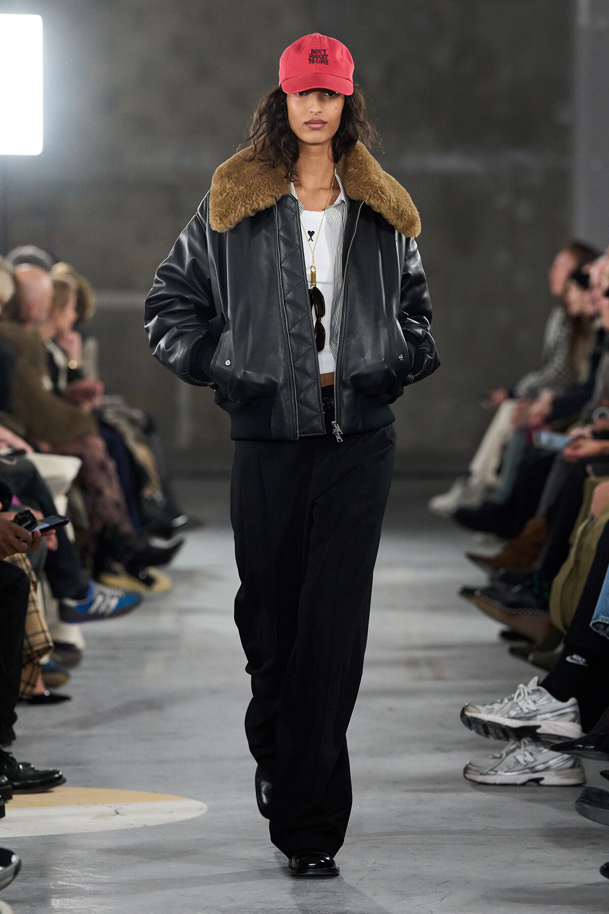 Runway model in a black leather bomber with brown shearling collar, white top, wide-leg black pants, and a red baseball cap.
