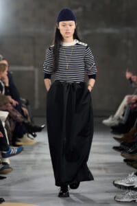 Model walking a runway in a navy and white striped sweater with a long black, gathered skirt and a navy knit cap.