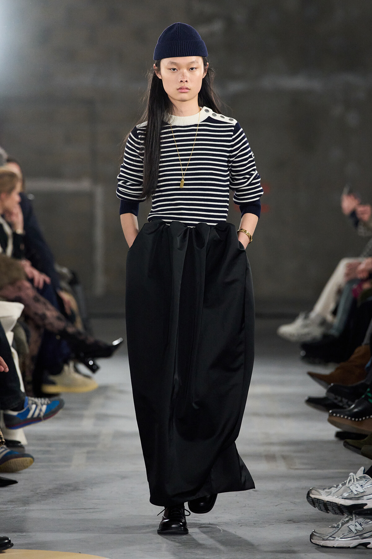 Model walking a runway in a navy and white striped sweater with a long black, gathered skirt and a navy knit cap.