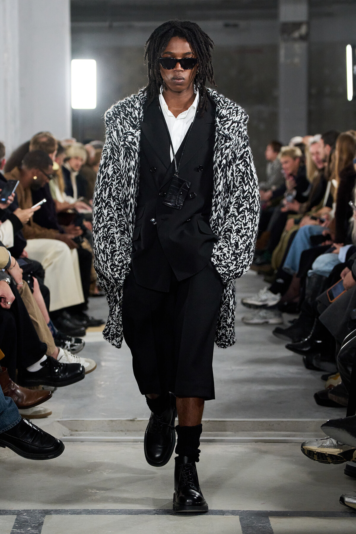 Model walking a runway in a black double-breasted suit beneath a chunky black-and-white knit coat, with dark sunglasses.