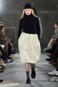 Model walking the runway in a black knit turtleneck and ivory gathered midi skirt, with a black beanie and gray socks paired with black shoes.