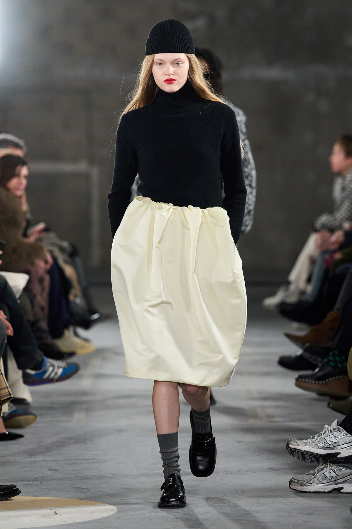 Model walking the runway in a black knit turtleneck and ivory gathered midi skirt, with a black beanie and gray socks paired with black shoes.