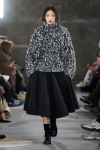Model walks the runway in a black-and-white chunky knit turtleneck sweater and a full black midi skirt; audience visible along the sides.
