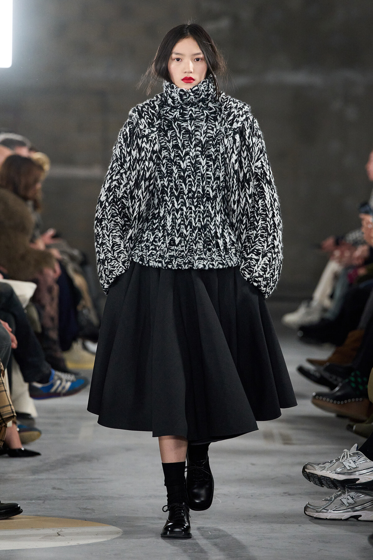 Model walks the runway in a black-and-white chunky knit turtleneck sweater and a full black midi skirt; audience visible along the sides.