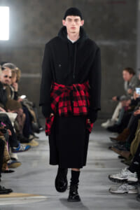 Male model on a runway wearing a long black coat over a hoodie, with a red plaid shirt tied at the waist and black boots.