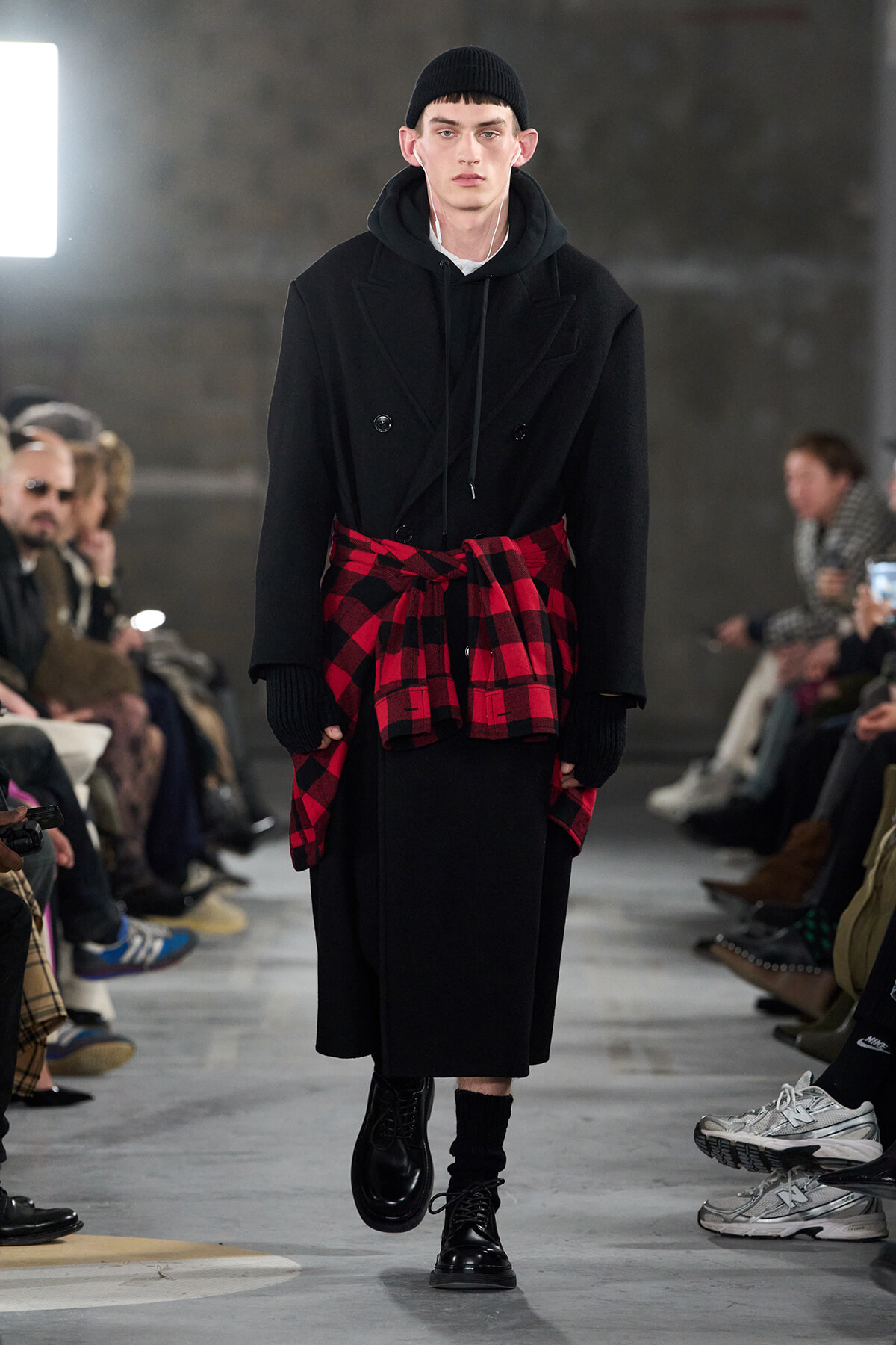 Male model on a runway wearing a long black coat over a hoodie, with a red plaid shirt tied at the waist and black boots.