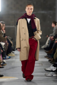 Model on a runway wearing a beige double-breasted coat, navy-black shirt, red wide-leg pants, and a navy/red striped scarf tied around the neck.