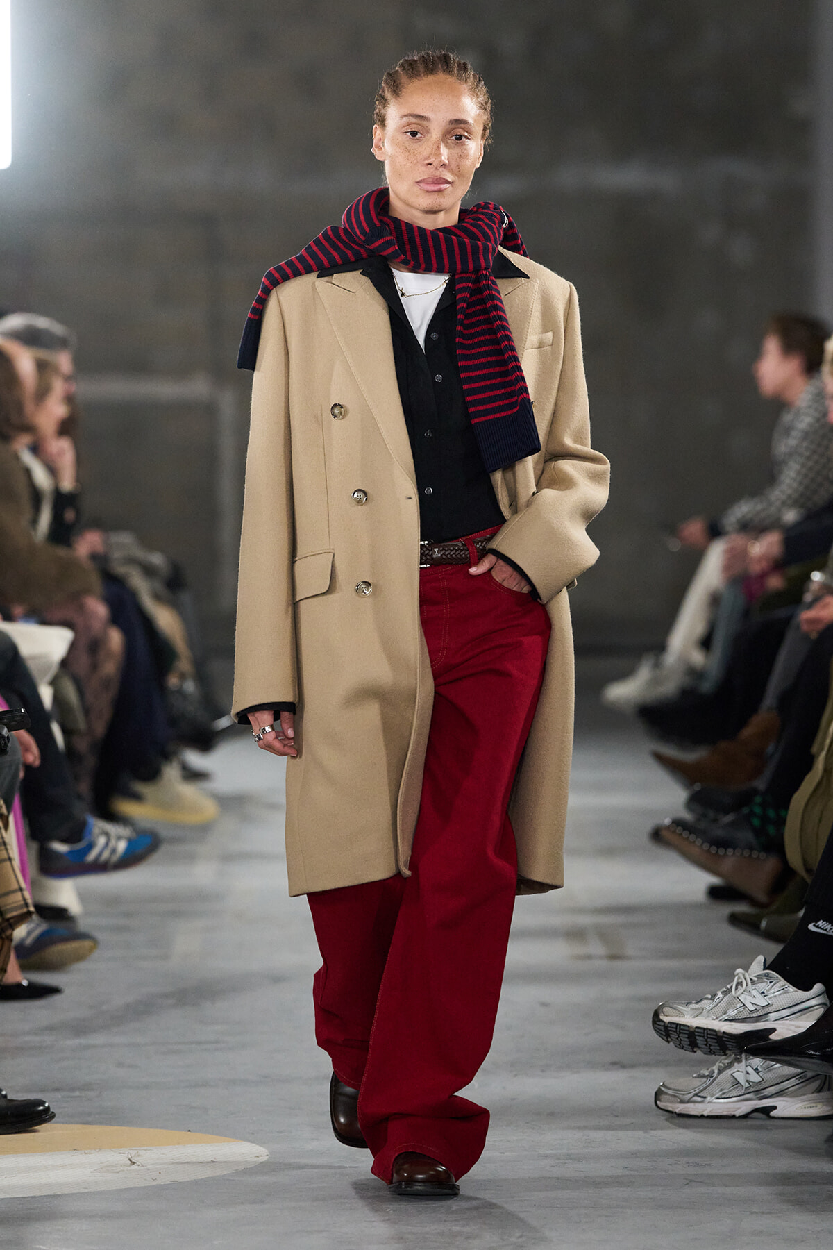 Model on a runway wearing a beige double-breasted coat, navy-black shirt, red wide-leg pants, and a navy/red striped scarf tied around the neck.