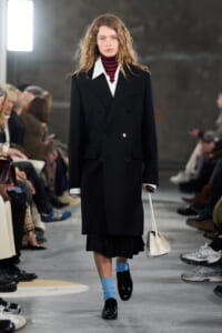 Runway model wearing a long black double-breasted coat over a white shirt and red striped turtleneck, bright blue socks, black patent shoes, and a small white handbag.