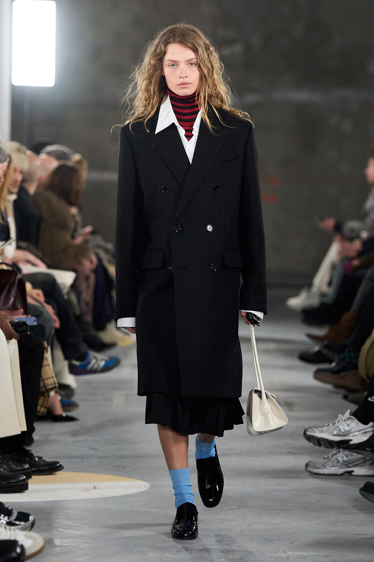 Runway model wearing a long black double-breasted coat over a white shirt and red striped turtleneck, bright blue socks, black patent shoes, and a small white handbag.