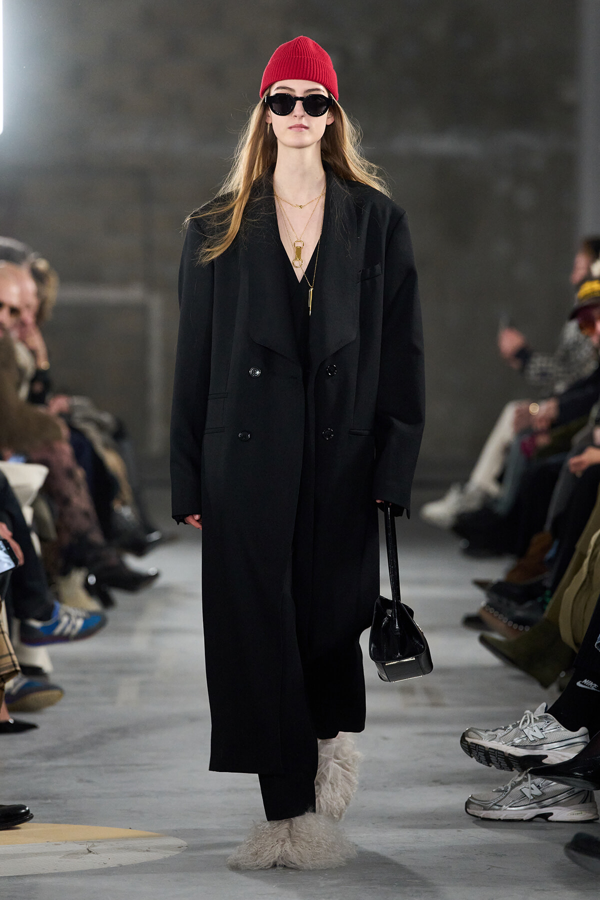 Runway model in an oversized black coat, red beanie, sunglasses, gold necklaces, fuzzy boots, and a small black handbag.