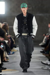 Male model walks a runway in a black-and-white varsity jacket, gray wide trousers, and a green cap, with sunglasses hanging from his sweater.